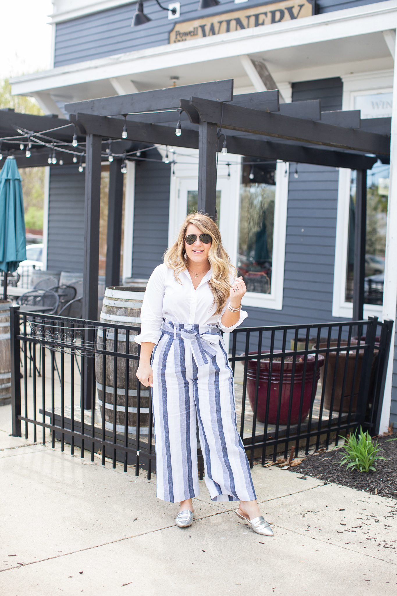 Blue Striped Paperbag Pants by popular North Carolina fashion blog, Coffee Beans and Bobby Pins: image of a woman standing outside a blue building with bistro lighting and wearing J. Crew Petite Point Sur paper-bag pant in faded indigo stripe, white Madewell Tie Front Shirt, Nordstrom BP. Ander 62mm Round Sunglasses, J. Crew Rainbow raffia fringe disc earrings, Target Women's Junebug Mules by A New Day, and Madewell Coin Necklace Set.
