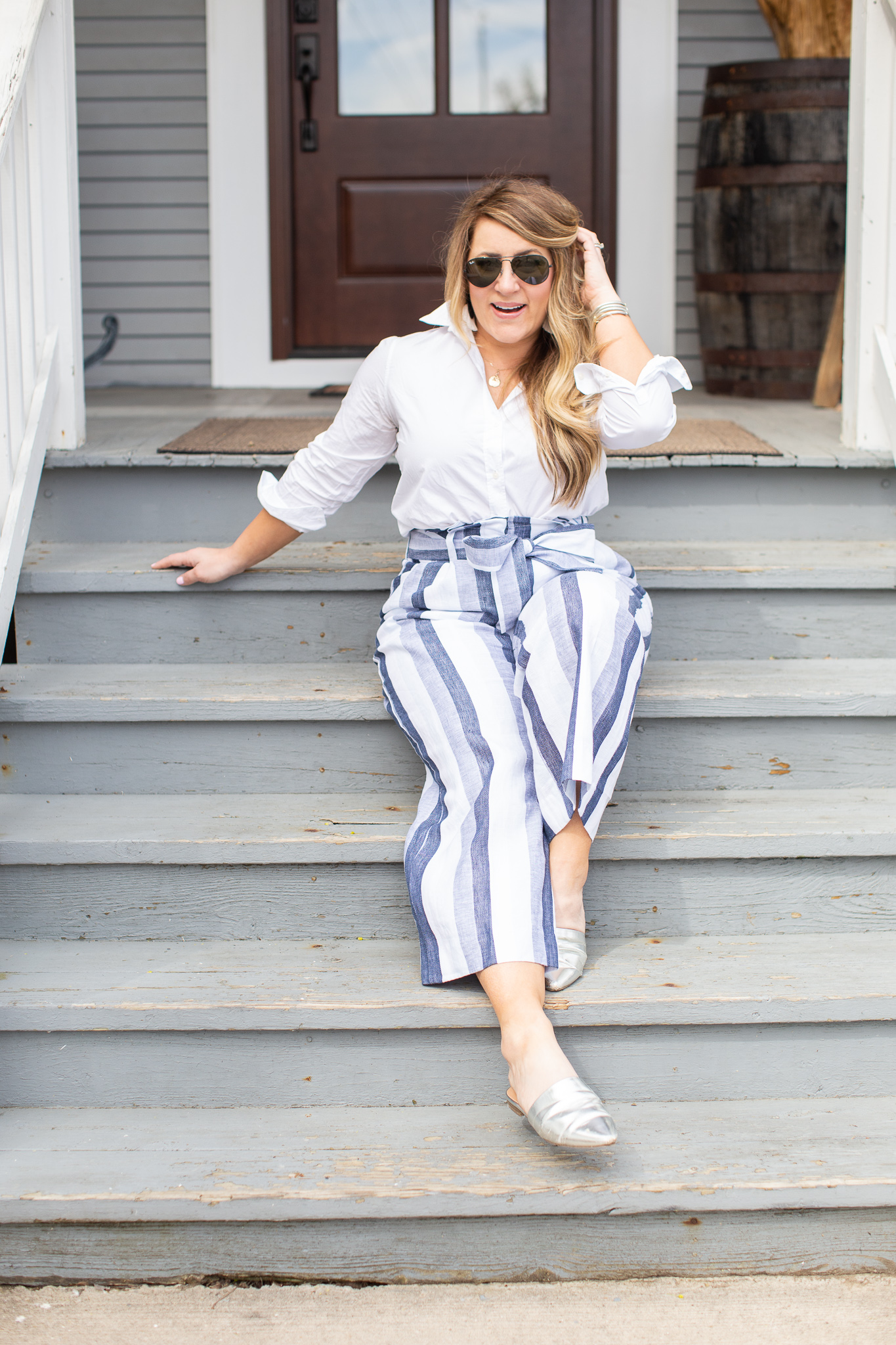 Blue Striped Paperbag Pants by popular North Carolina fashion blog, Coffee Beans and Bobby Pins: image of a woman sitting outside on the steps of a blue building and wearing J. Crew Petite Point Sur paper-bag pant in faded indigo stripe, white Madewell Tie Front Shirt, Nordstrom BP. Ander 62mm Round Sunglasses, J. Crew Rainbow raffia fringe disc earrings, Target Women's Junebug Mules by A New Day, and Madewell Coin Necklace Set.