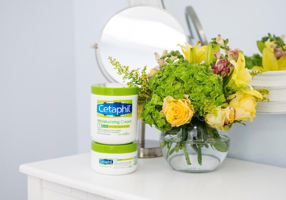 Summer Skin Care Tips with Cetaphil by popular North Carolina beauty blog, Coffee Beans and Bobby Pins: image of two Cetaphil Moisturizing Cream containers stacked on top of each other on a white vanity next to a vase of flowers.