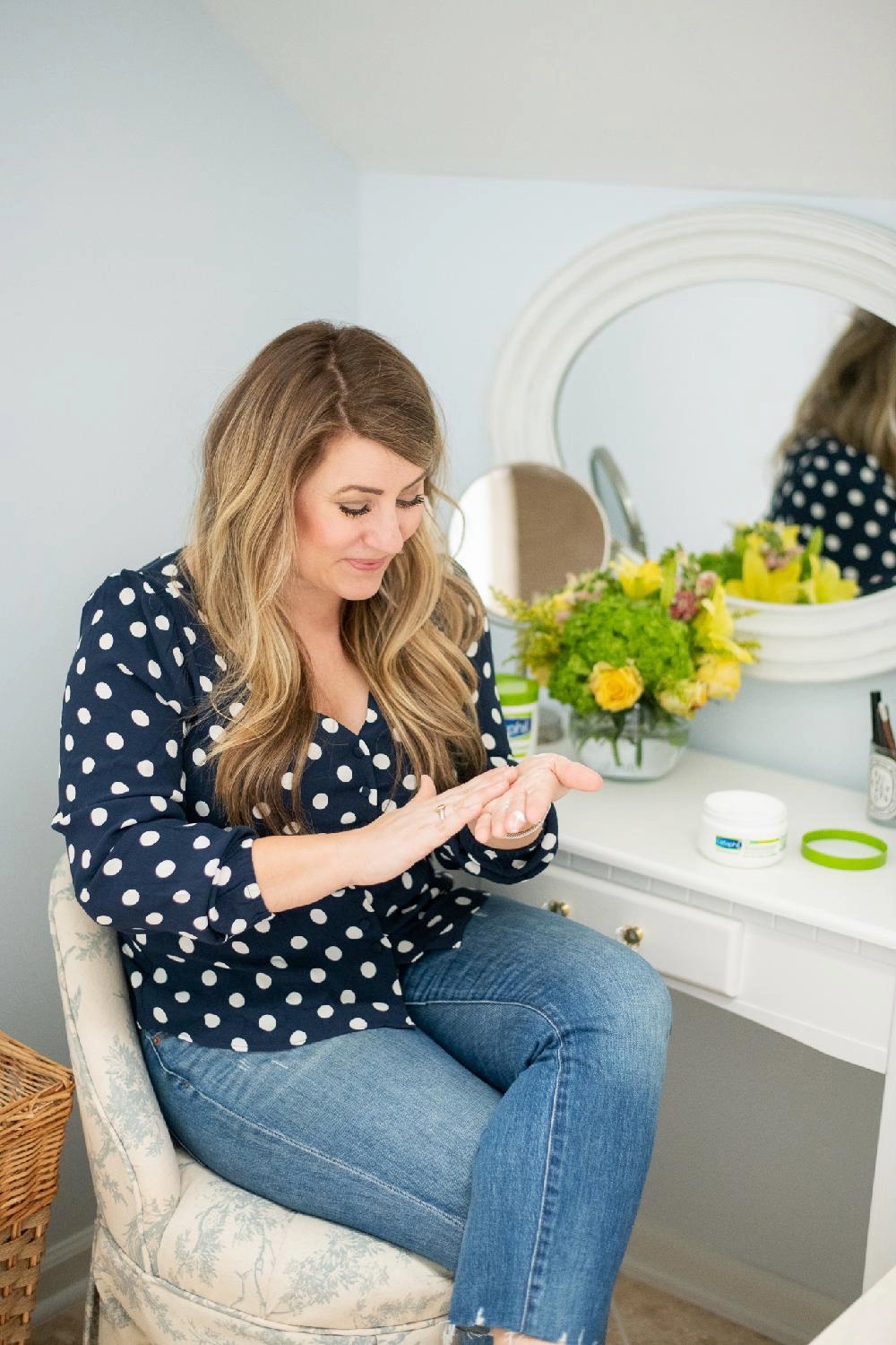 Summer Skin Care Tips with Cetaphil by popular North Carolina beauty blog, Coffee Beans and Bobby Pins: image of woman sitting down at her vanity and applying Cetaphil Moisturizing Cream to her hands.