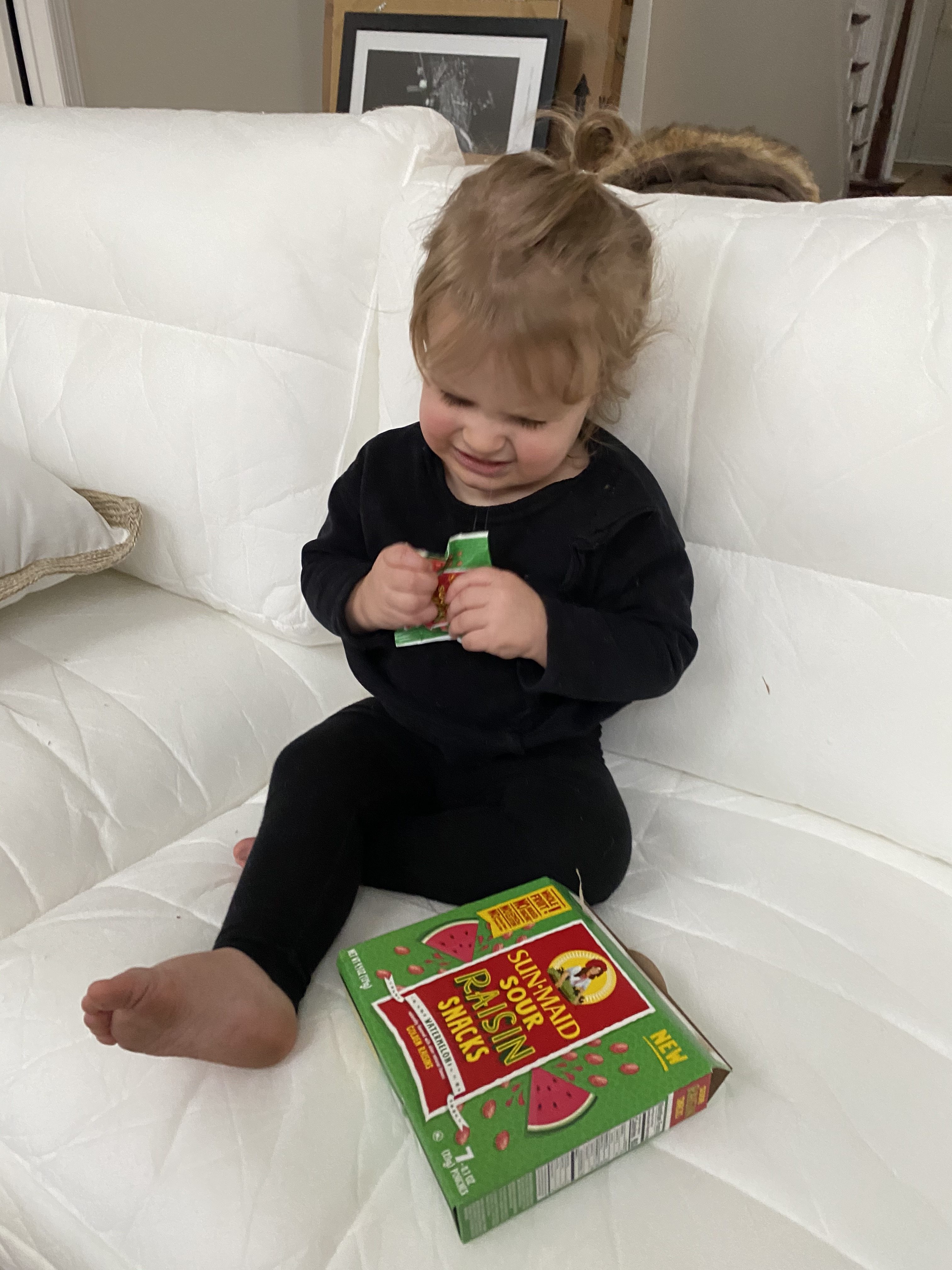 Quick Snacks for Toddlers by popular Ohio mom blog, Coffee Beans and Bobby Pins: image of a little girl sitting on a white couch and eating Sun-Maid Sour Raisin snacks. 