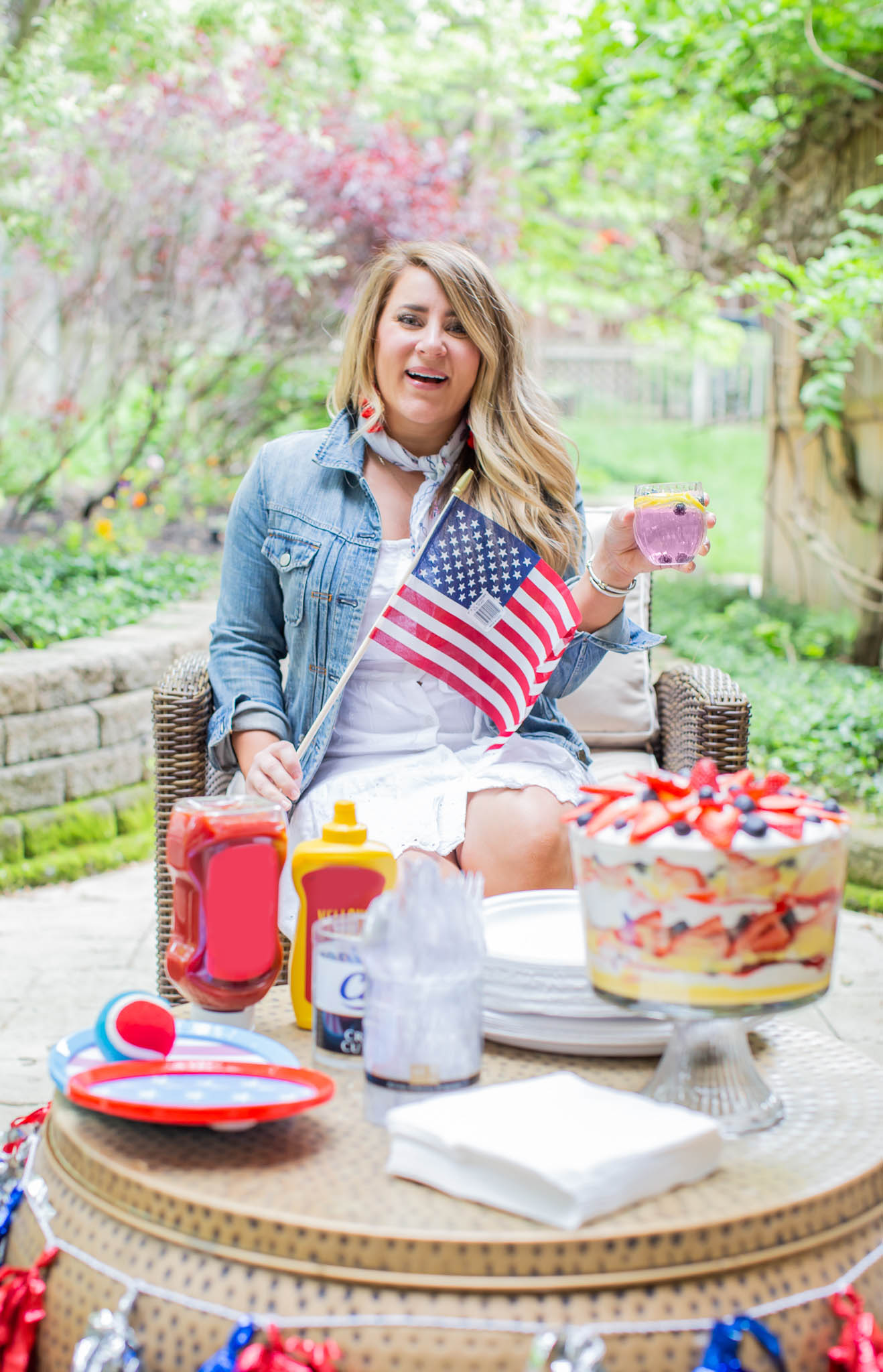 How to Throw the Ultimate (and Easy) 4th of July Party by popular North Carolina blog, Coffee Beans and Bobby Pins: image of woman sitting outside and holding an american flag with a table that has white chinet plates, white chinet napkins, chinet cut crystal cutlery, a red white and blue tennis ball, 4ht of July paper plates, ketchup, mustard, 4th of July decorations and a berry trifle on it. 