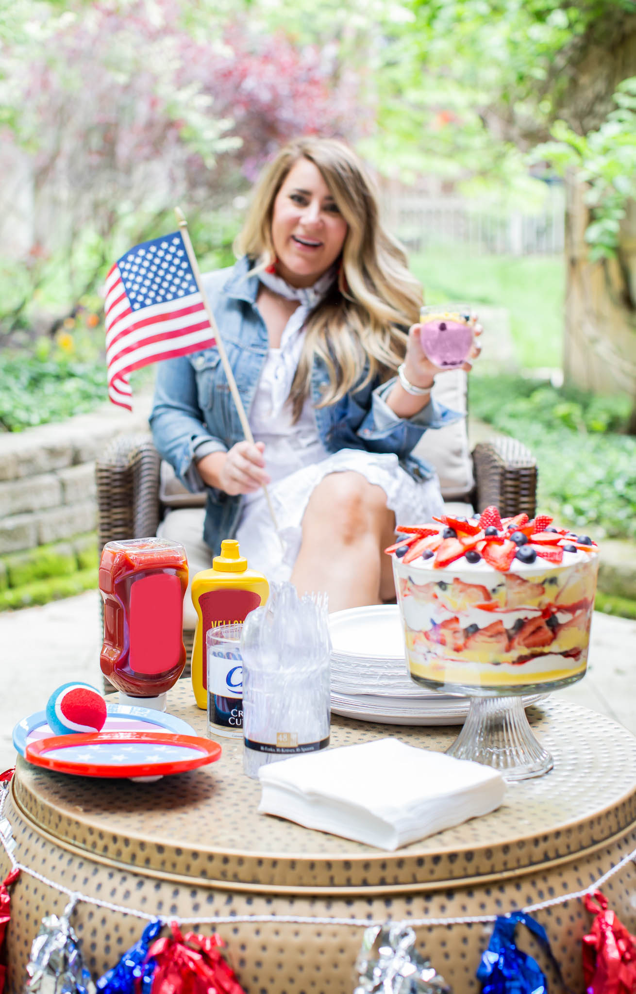 How to Throw the Ultimate (and Easy) 4th of July Party by popular North Carolina blog, Coffee Beans and Bobby Pins: image of woman sitting outside and holding an american flag with a table that has white chinet plates, white chinet napkins, chinet cut crystal cutlery, a red white and blue tennis ball, 4ht of July paper plates, ketchup, mustard, 4th of July decorations and a berry trifle on it. 