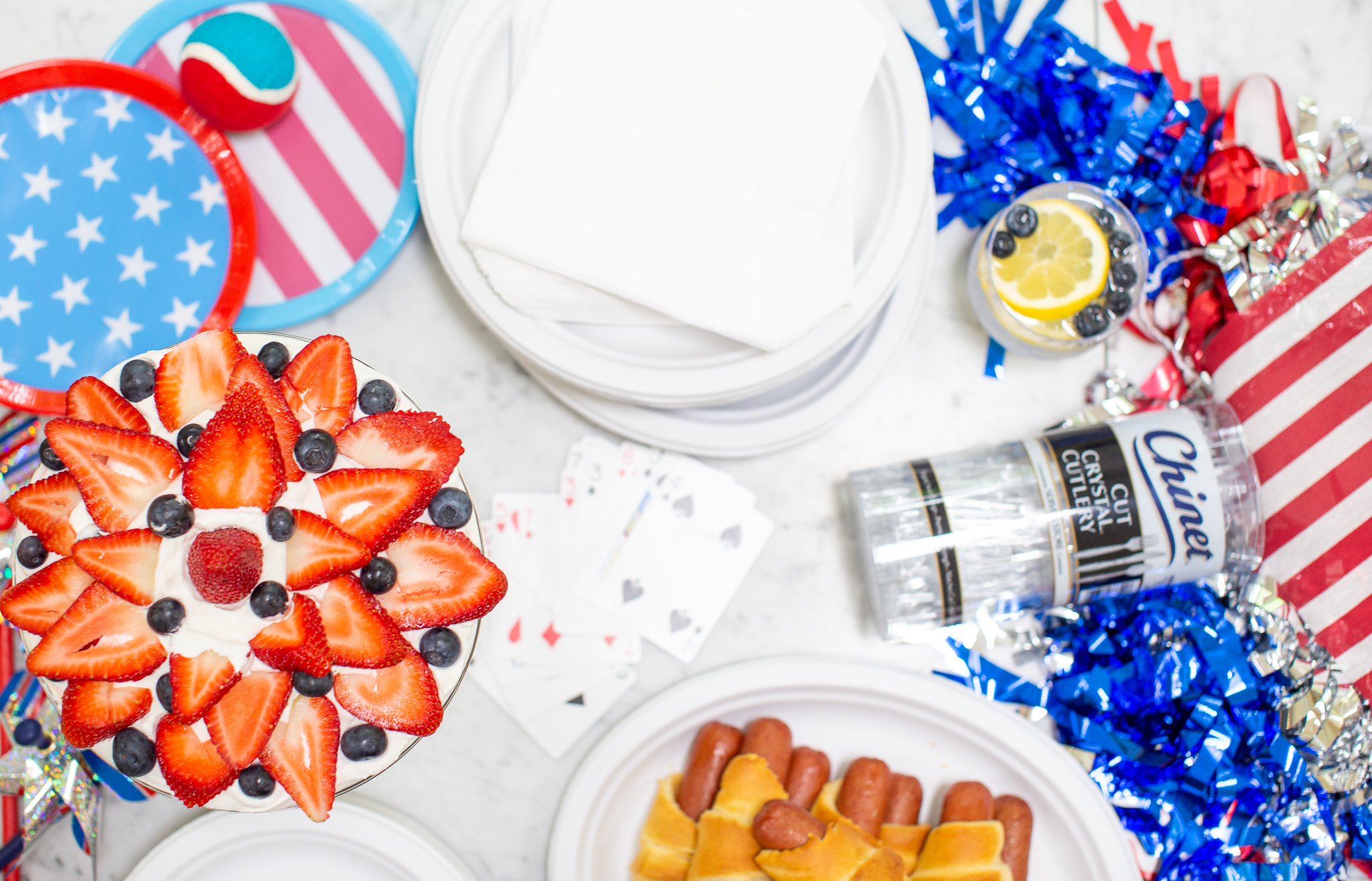How to Throw the Ultimate (and Easy) 4th of July Party by popular North Carolina blog, Coffee Beans and Bobby Pins: image of white chinet plates, white chinet napkins, chinet cut crystal cutlery, 4th of July paper plates, red white and blue tennis ball, hot dogs, face cards, 4th of July decorations and a berry trifle. 