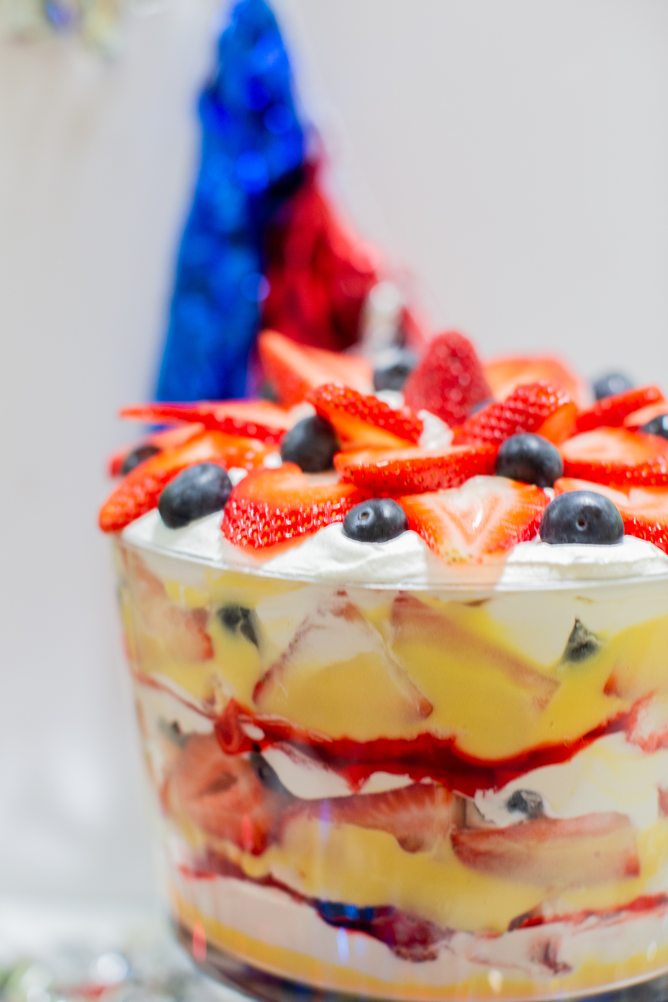 Three Berry Vanilla Pudding Trifle Recipe (the Perfect Summertime Dessert) by popular lifestyle blog, Coffee Beans and Bobby Pins: image of a three berry vanilla pudding trifle in a trifle dish with a red and blue tassel banner hanging in the background.