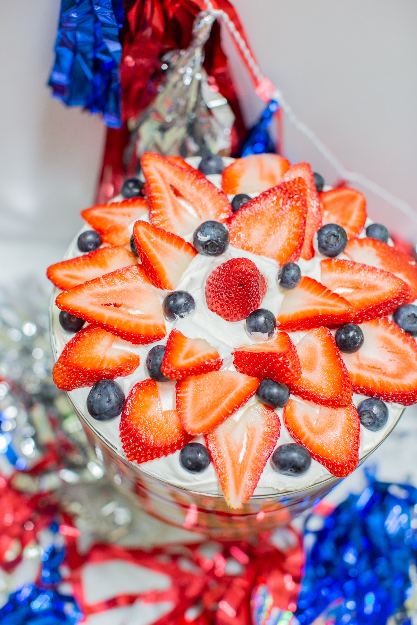 Three Berry Vanilla Pudding Trifle Recipe (the Perfect Summertime Dessert) by popular lifestyle blog, Coffee Beans and Bobby Pins: image of a three berry vanilla pudding trifle in a trifle dish with a red and blue tassel banner hanging in the background.