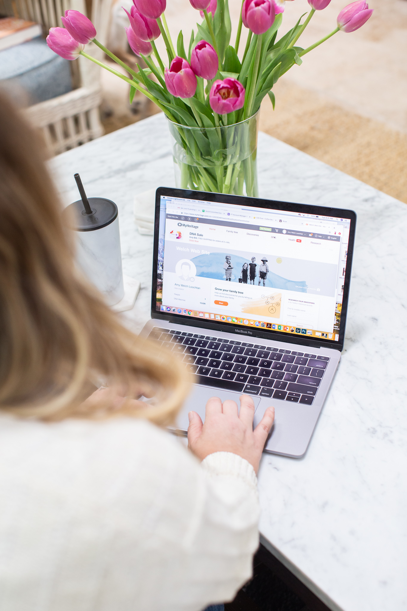 MyHeritage DNA Results by popular North Carolina fashion blog, Coffee Beans and Bobby Pins: image of a woman looking at her opened laptop that is resting on her marble top coffee table next to a vase of pink tulips.