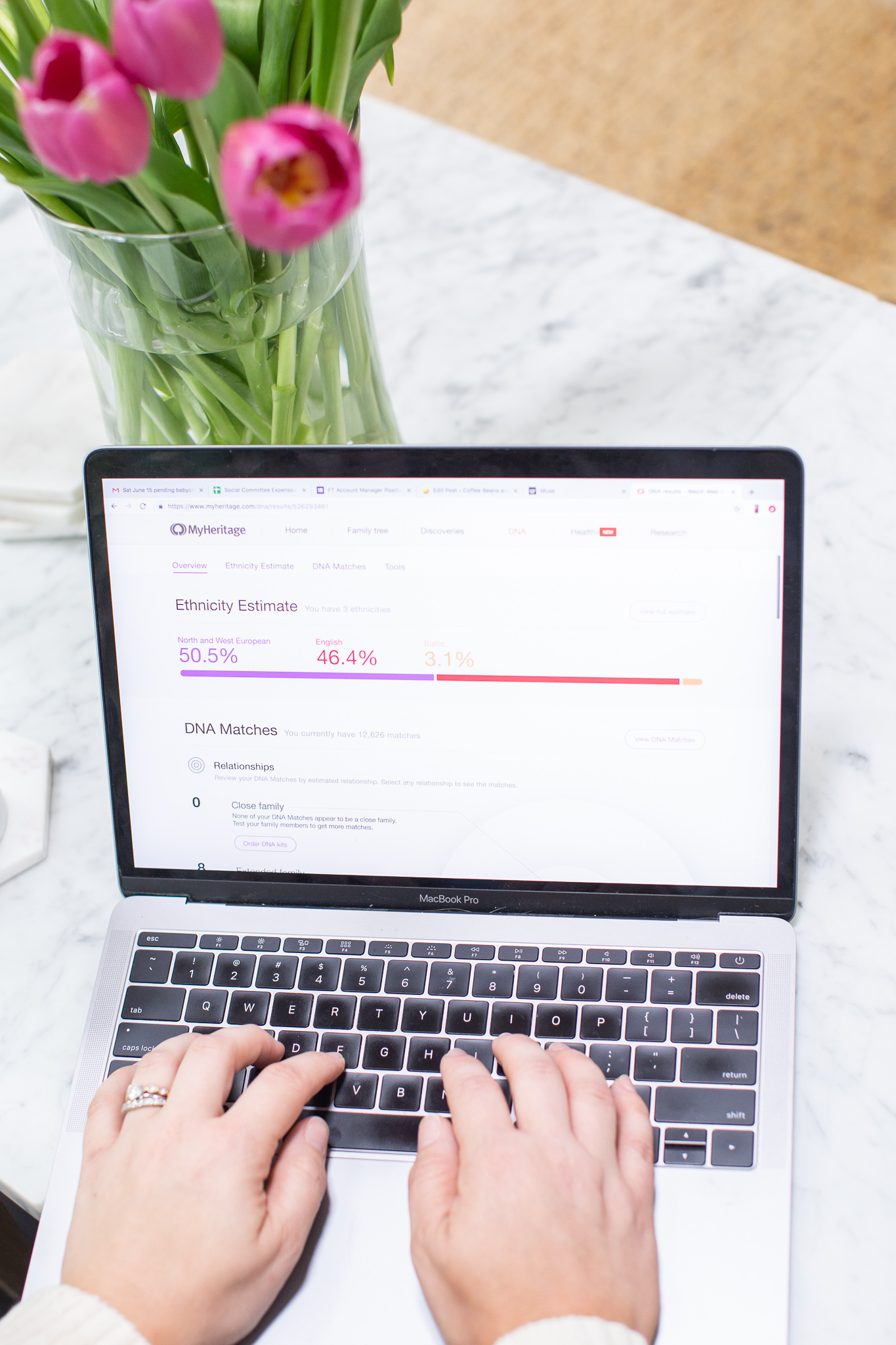 MyHeritage DNA Results by popular North Carolina fashion blog, Coffee Beans and Bobby Pins: image of a woman looking at her opened laptop that is resting on her marble top coffee table next to a vase of pink tulips.