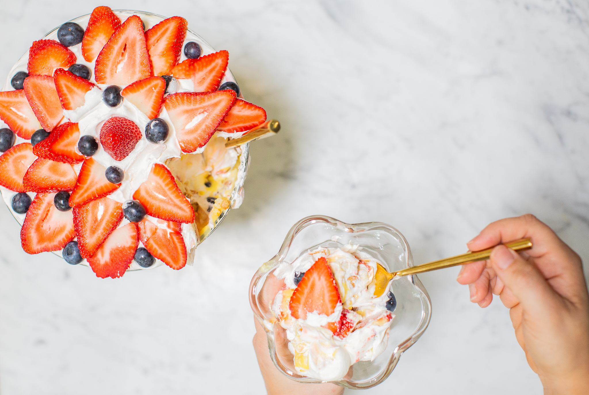 Three Berry Vanilla Pudding Trifle Recipe (the Perfect Summertime Dessert) by popular lifestyle blog, Coffee Beans and Bobby Pins: flatlay image of a three berry vanilla pudding trifle and a hand holding a clear glass desert dish with a portion of the trifle in it.