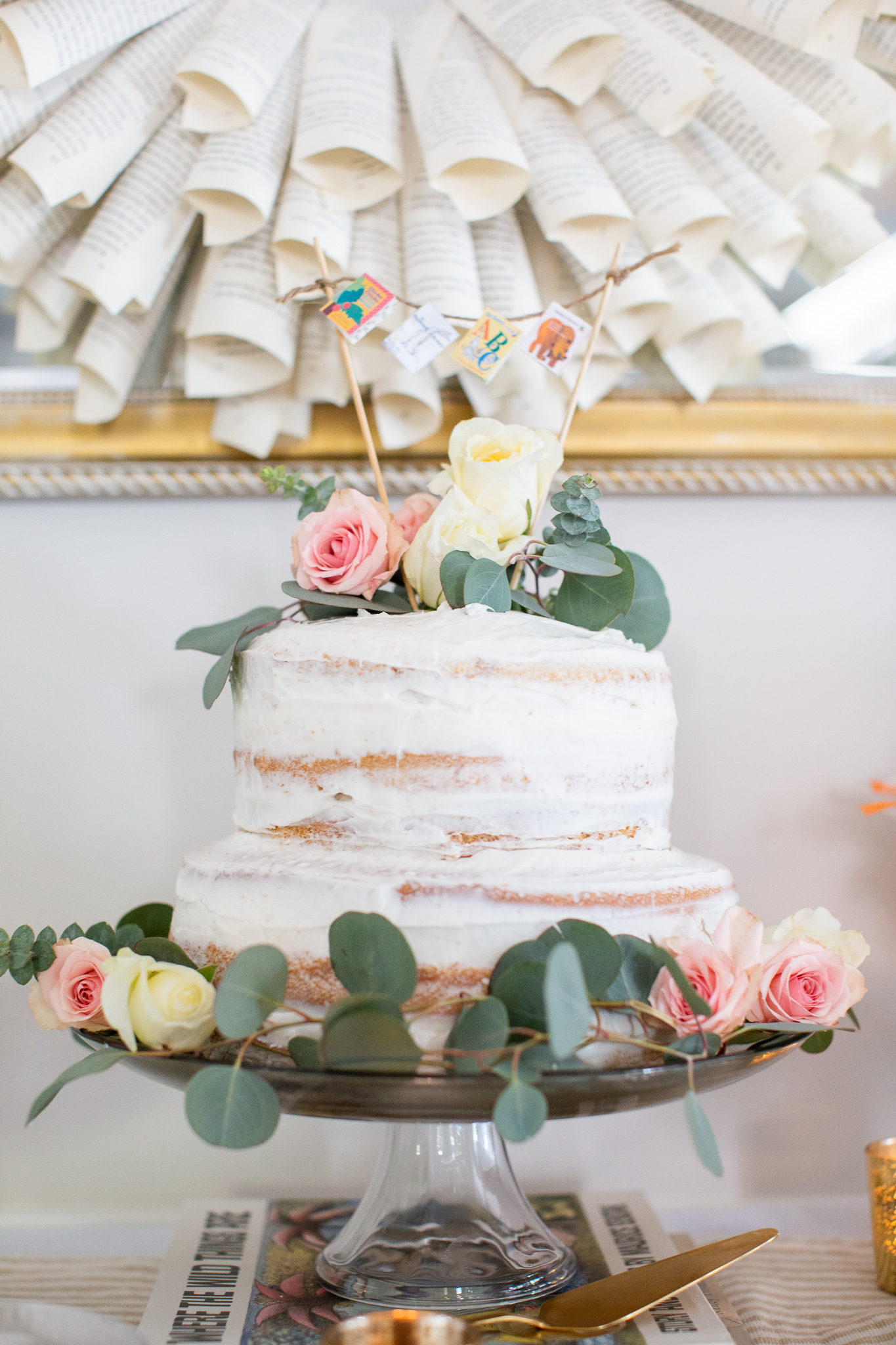  Story Book Baby Shower Ideas by popular North Carolina life and style blog, Coffee Beans and Bobby Pins: image of a two tier cake with a picture book pennant banner cake topper and fresh flowers. 