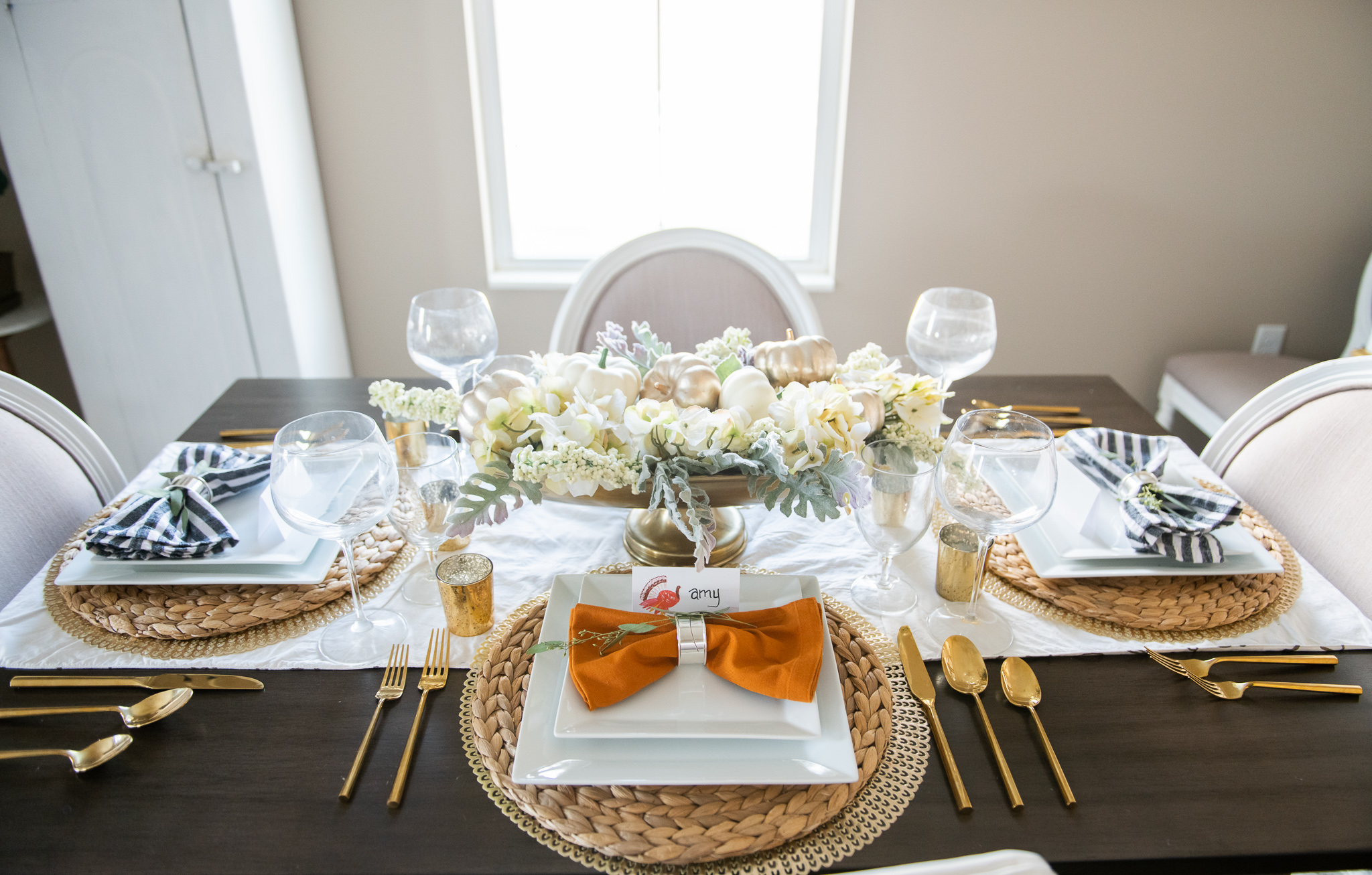 Thanksgiving Tablescape Ideas (and a small update) by popular North Carolina lifestyle blog, Coffee Beans and Bobby Pins: image of a table set with wicker placemats, gold silverware, wine glasses, white floral centerpiece, white table runner, cloth napkins, and white square plates. 