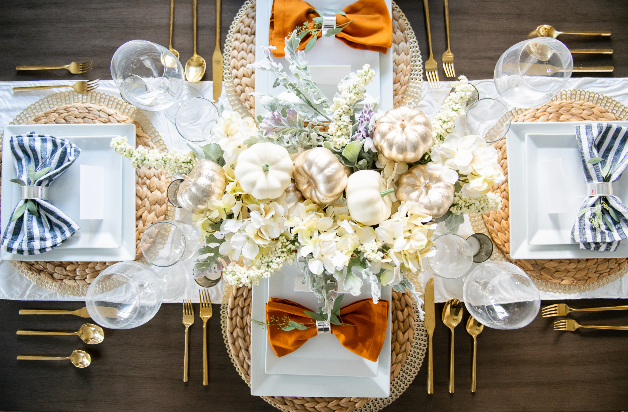 Thanksgiving Tablescape Ideas (and a small update) by popular North Carolina lifestyle blog, Coffee Beans and Bobby Pins: image of a table set with wicker placemats, gold silverware, wine glasses, white floral centerpiece, white table runner, cloth napkins, and white square plates. 