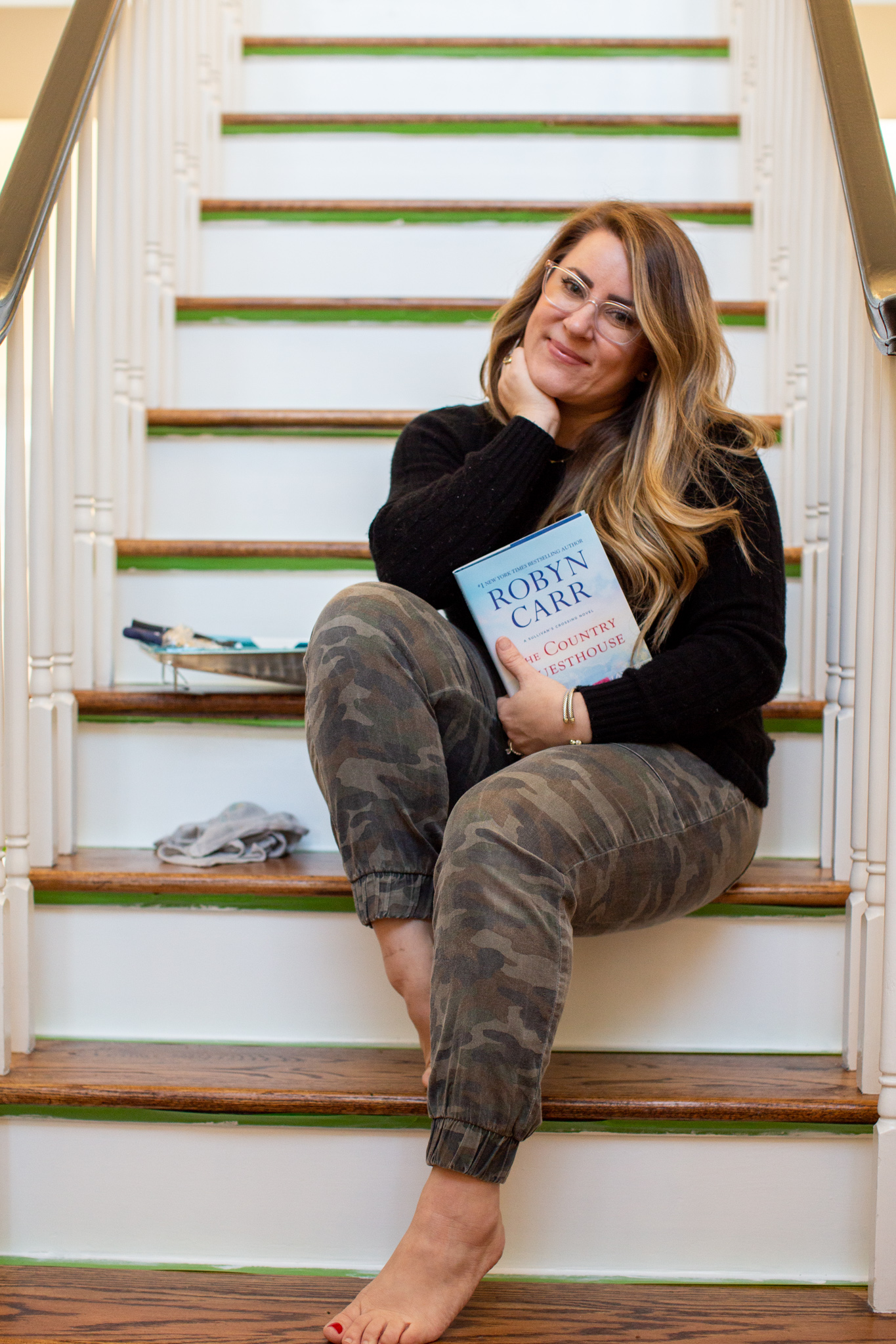 Hey, hey 2020. Read this book. by popular Ohio lifestyle blog, Coffee Beans and Bobby Pins: image of a woman holding the book The Country Guesthouse and sitting on a painted staircase.