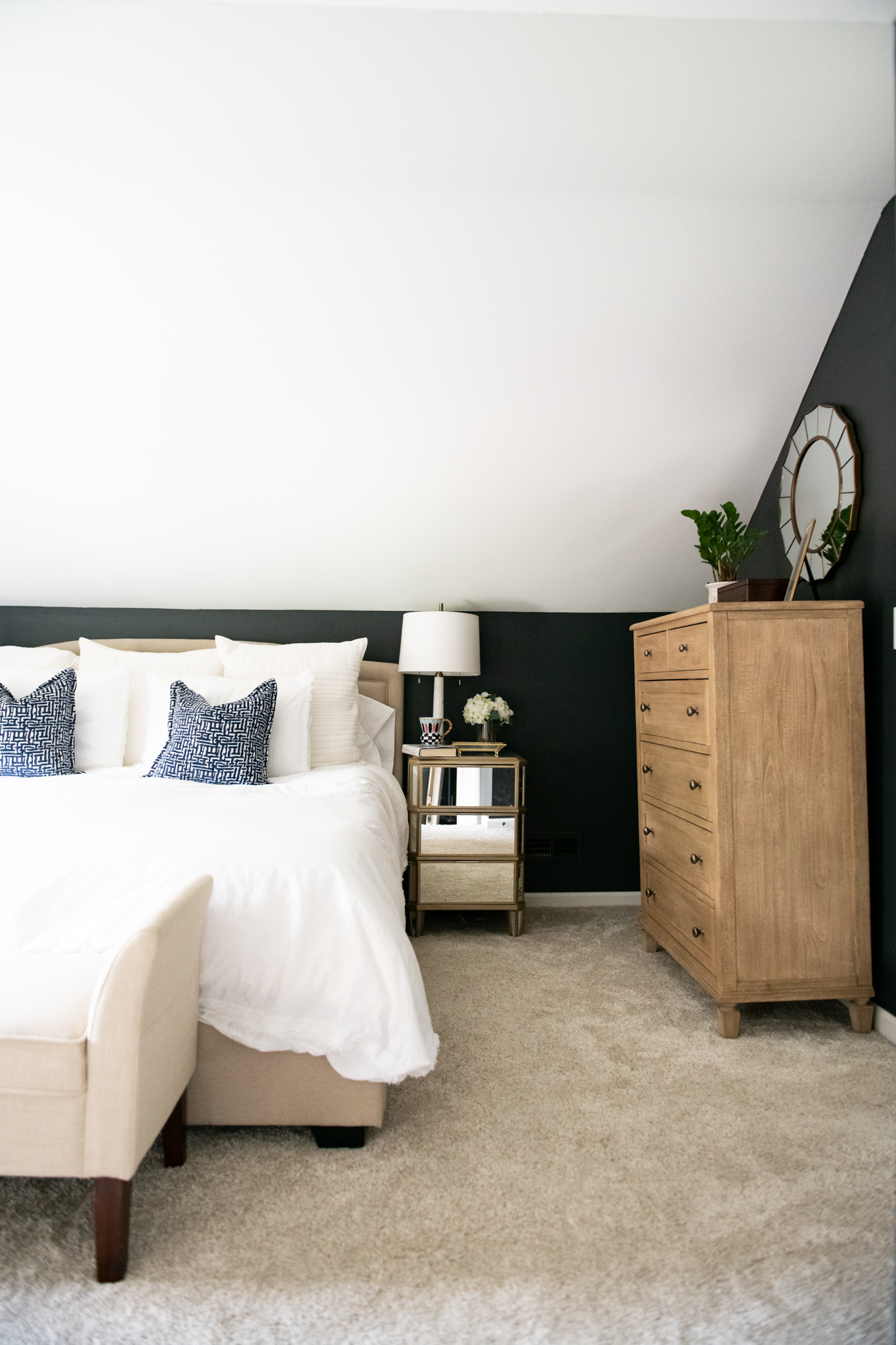 Master Bedroom Remodel Ideas by popular Ohio life and style blog, Coffee Beans and Bobby Pins: image of a master bedroom with a wood bead chandelier, black walls, mirrored night stands, light wood dresser, round mirror, king size bed with white linens and a cream fabric bench at the foot of the bed.