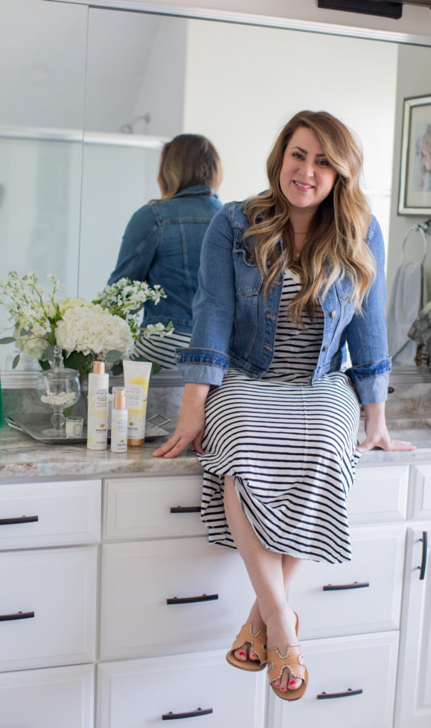 Pantene Fortifying Damage Repair Collection Reviewed by popular Ohio beauty blog, Coffee Beans and Bobby Pins: image of a woman wearing a Evereve Z Supply Reverie Stripe Dress, Evereve Steven Greece Sandal, and Evereve Kut From The Kloth Amelia Denim Jacket and sitting on her bathroom counter next to some Pantene castor oil hair products.