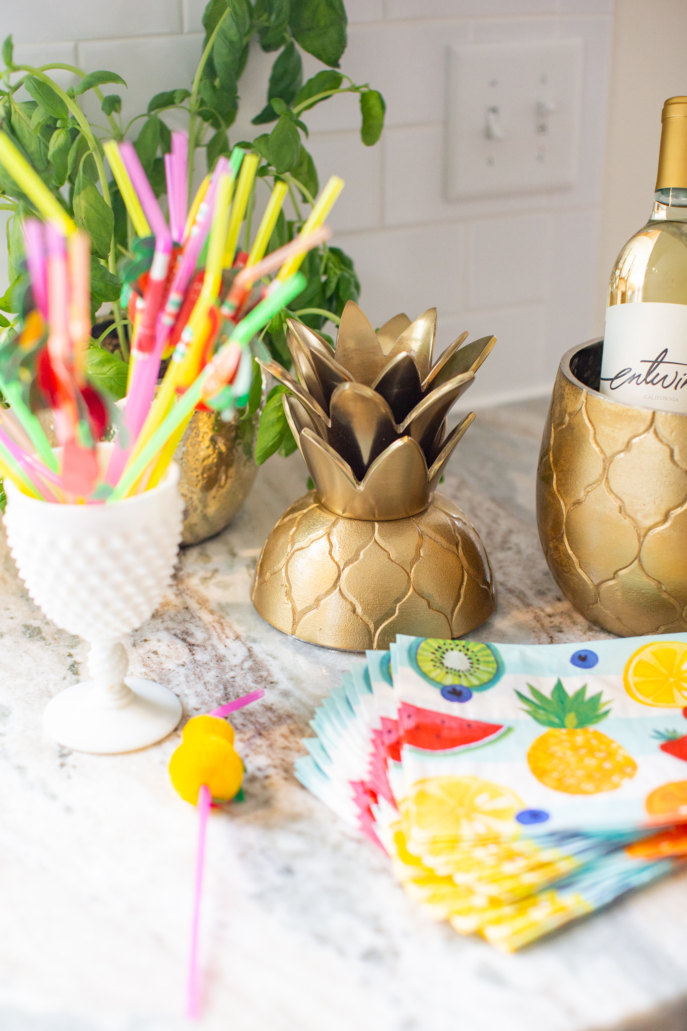 2nd Birthday Party Ideas by popular Ohio lifestyle blog, Coffee Beans and Bobby Pins: image of a white milk glass cup holding colored straws, fruit print napkins, and a gold pineapple wine chiller. 
