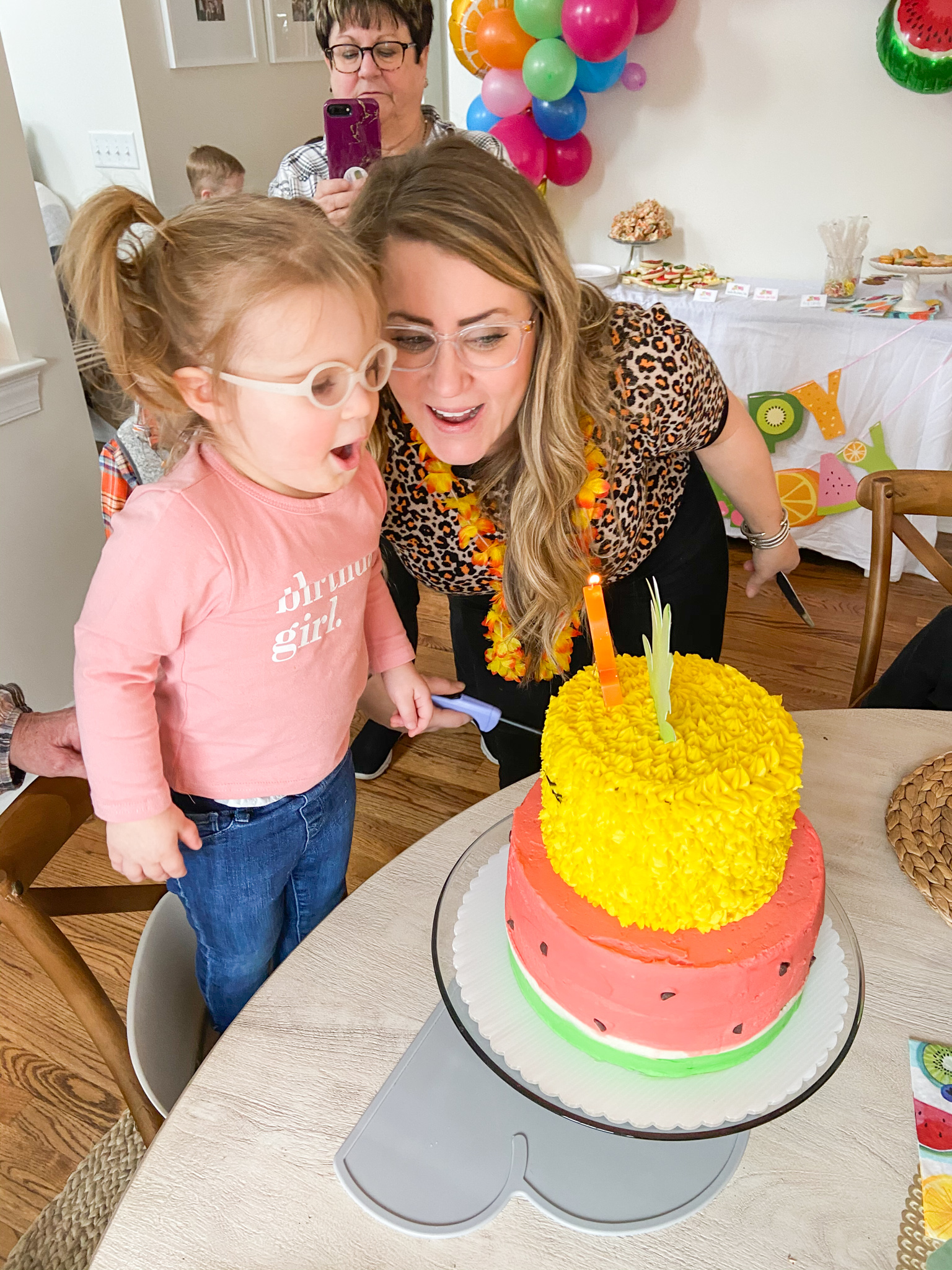 2nd Birthday Party Ideas by popular Ohio lifestyle blog, Coffee Beans and Bobby Pins: image of a mom standing next to her two year old daughter and helping her blow out a number 2 candle on her 2 tier pineapple and watermelon decorated cake.