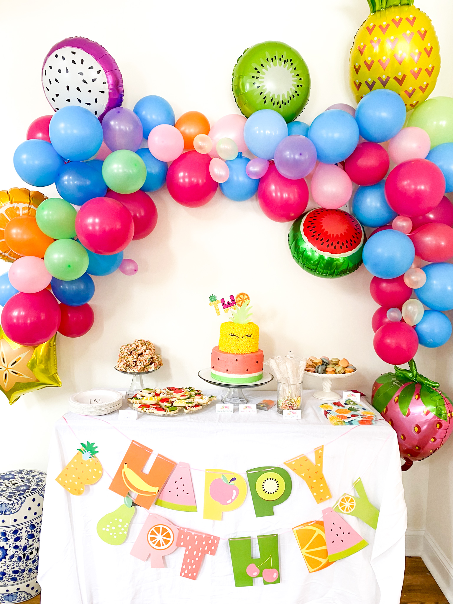 2nd Birthday Party Ideas by popular Ohio lifestyle blog, Coffee Beans and Bobby Pins: image of a multi color balloon garland with Mylar fruit balloons hanging above a dessert table containing two tier pineapple and watermelon decorated cake, rainbow colored Rice Krispy treats,, white paper plates, a white table cloth, fruit print napkins, macaroons, and fruit shaped sugar cookies.  