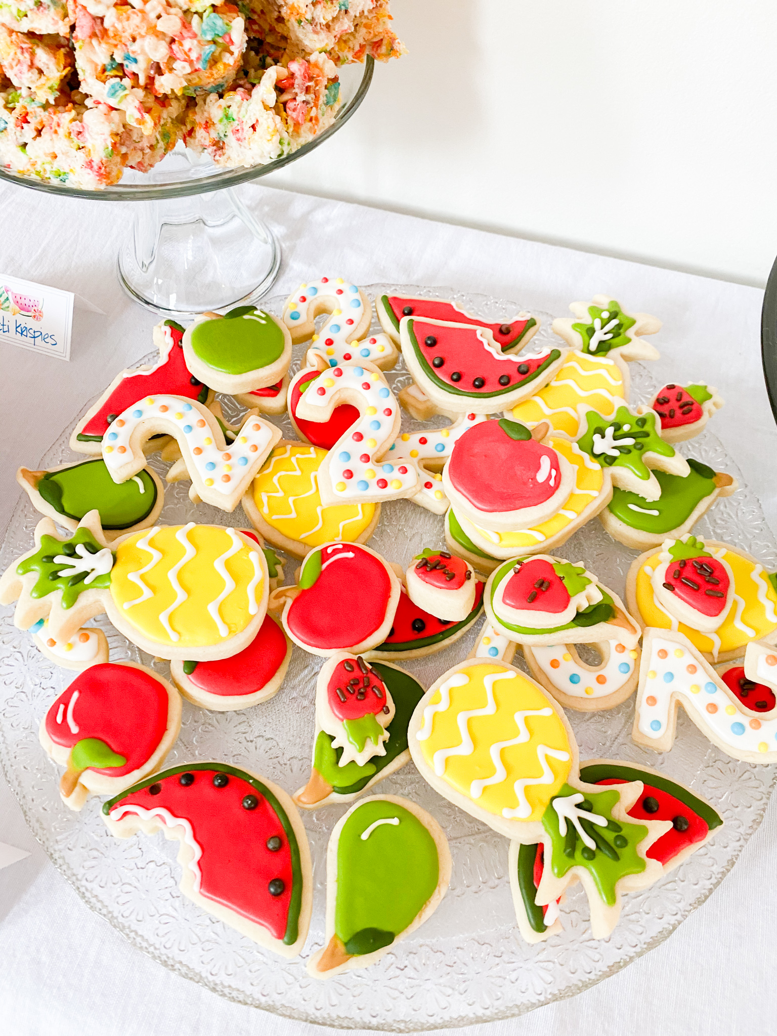 2nd Birthday Party Ideas by popular Ohio lifestyle blog, Coffee Beans and Bobby Pins: image of a dessert table containing rainbow colored Rice Krispy treats and sugar cookies decorated to look like apples, watermelon slices, strawberries, pineapple, and the number 2.  