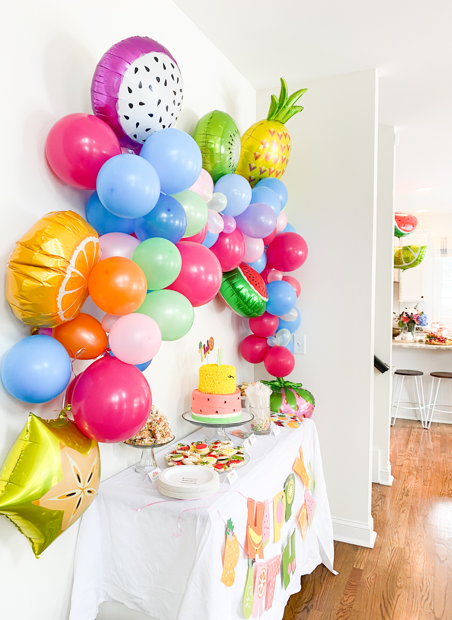 2nd Birthday Party Ideas by popular Ohio lifestyle blog, Coffee Beans and Bobby Pins: image of a multi color balloon garland with Mylar fruit balloons hanging above a dessert table containing two tier pineapple and watermelon decorated cake, rainbow colored Rice Krispy treats,, white paper plates, a white table cloth, and sugar cookies.  