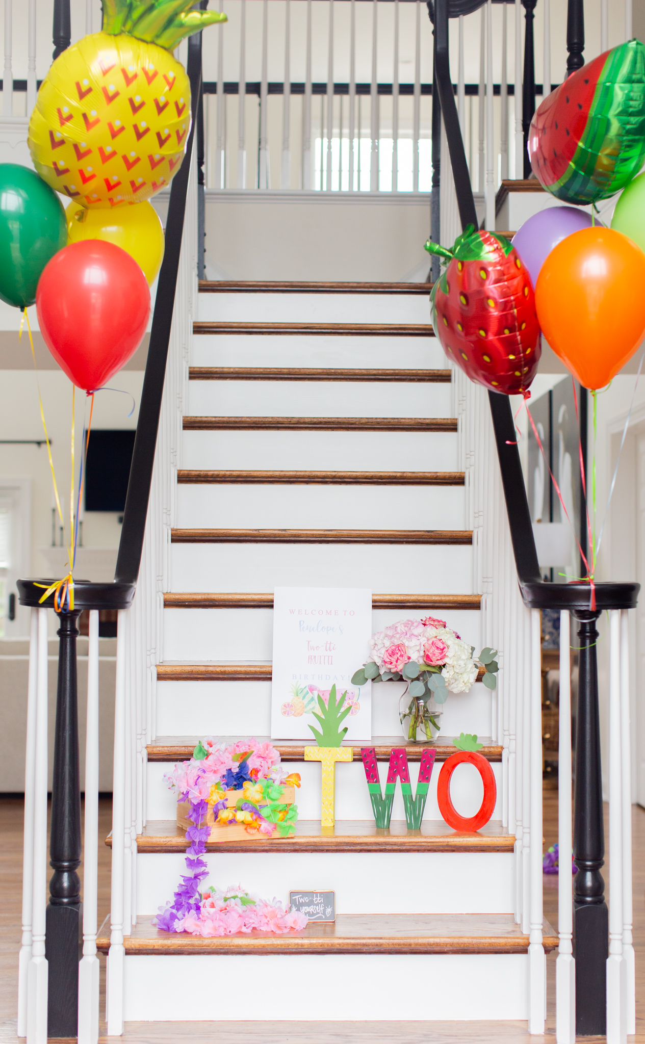 2nd Birthday Party Ideas by popular Ohio lifestyle blog, Coffee Beans and Bobby Pins: image of a staircase decorated with helium filled Mylar fruit balloons, regular helium filled balloons, fruit painted letters that spell out the number two, flower leis, and a floral arrangement of pink and white flowers.  