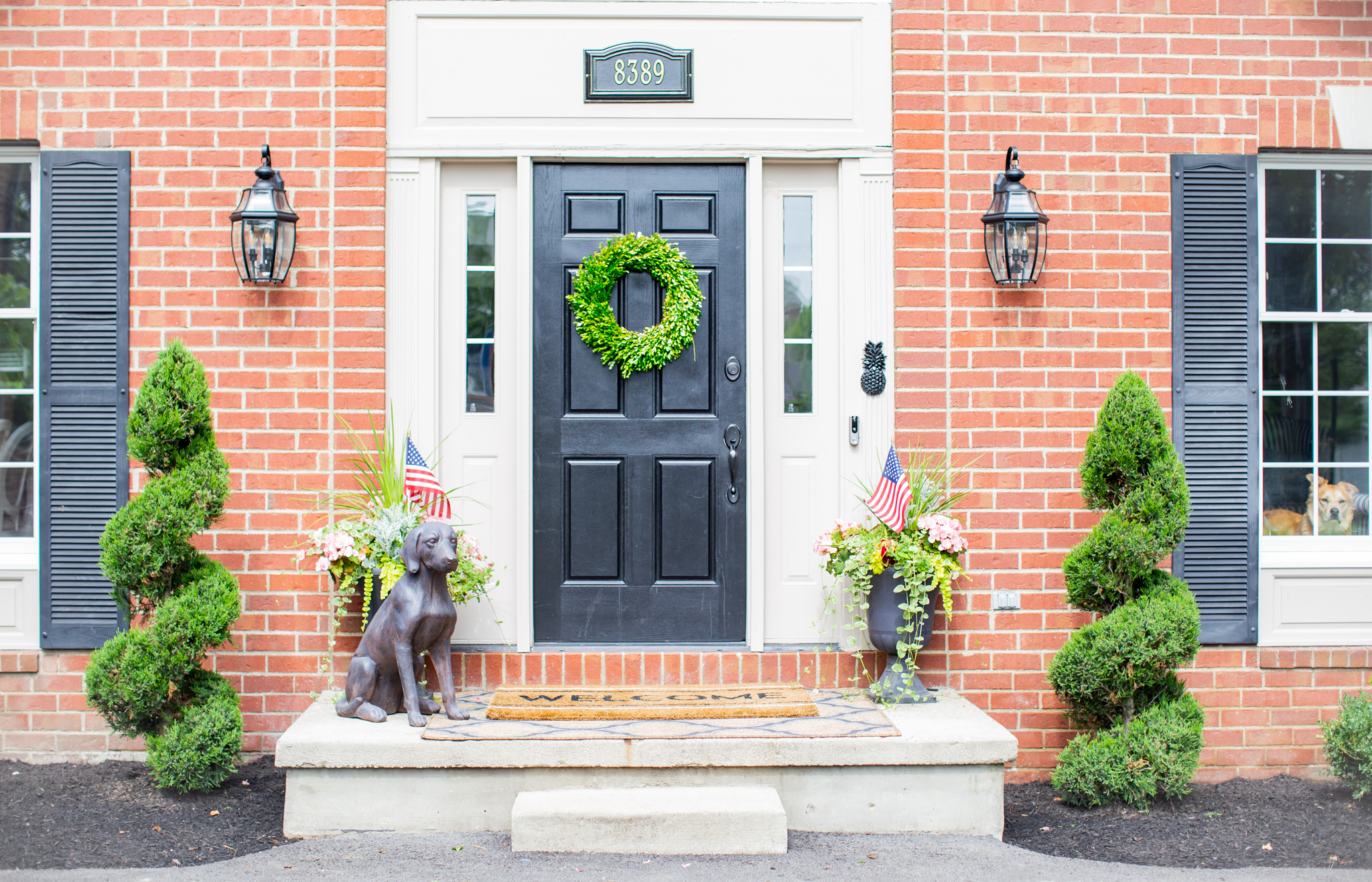 Front Porch by popular Ohio life and style blog, Coffee Beans and Bobby Pins: image of the front of a house decorated with American Flag bunting, Grandin Road Labrador Garden Statue, Grandin Road Hypnos Quatrefoil Outdoor Rug, Grandin Road Welcome Stripe Coir Door Mat, Grandin Road Sidney Urn/Planter, Grandin Road Soft Vine Wreath, Grandin Road Large Cambridge Single Line Wall Address Plaque, Grandin Road Devon Self-watering Window Box Planter, and Grandin Road Devon Tapered Planter.