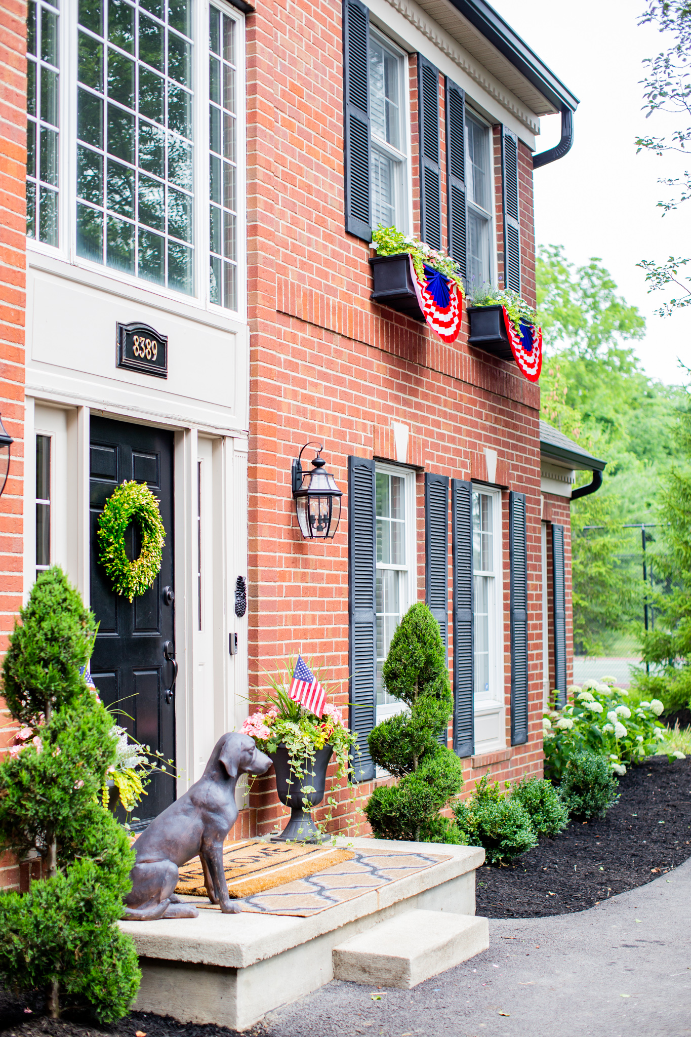 Front Porch by popular Ohio life and style blog, Coffee Beans and Bobby Pins: image of the front of a house decorated with American Flag bunting, Grandin Road Labrador Garden Statue, Grandin Road Hypnos Quatrefoil Outdoor Rug, Grandin Road Welcome Stripe Coir Door Mat, Grandin Road Sidney Urn/Planter, Grandin Road Soft Vine Wreath, Grandin Road Large Cambridge Single Line Wall Address Plaque, Grandin Road Devon Self-watering Window Box Planter, and Grandin Road Devon Tapered Planter.