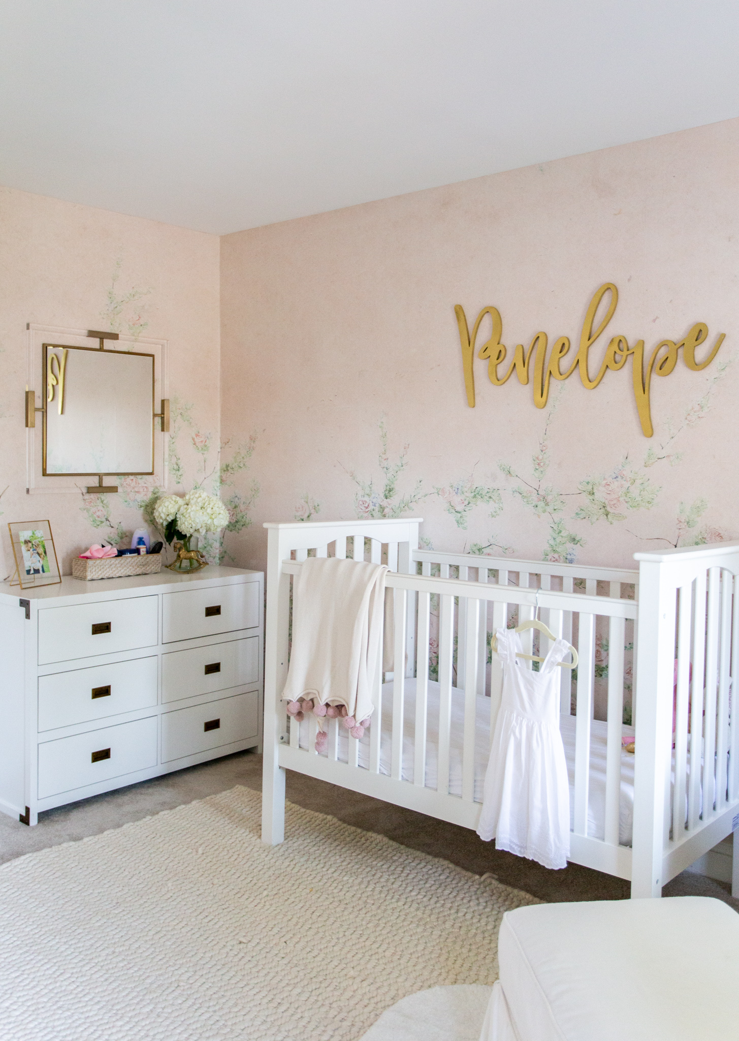 Floral Nursery by popular Ohio life and style blog, Coffee Beans and Bobby Pins: image of a nursery with Anewall Sweet Laurel Mural wallpaper, white armchair, white dresser, gold name sign, white crib, and a white ottoman. 
