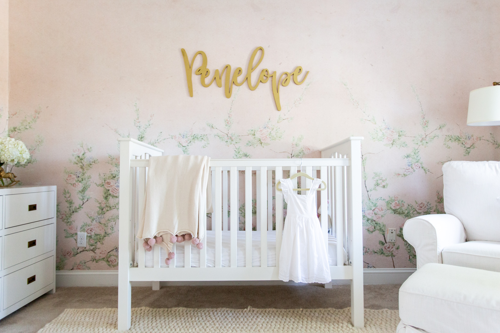 Floral Nursery by popular Ohio life and style blog, Coffee Beans and Bobby Pins: image of a nursery with Anewall Sweet Laurel Mural wallpaper, white armchair, white bookcase, gold name sign, gold wishbone decor, pink curtains and white crib.
