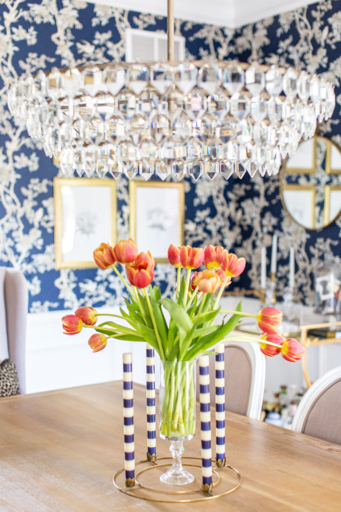 Blue Floral Wallpaper by popular Ohio life and style blog, Coffee Beans and Bobby Pins: image of a dining room with a crystal chandelier, blue floral wallpaper, wood floor, vase if red and yellow tulips, gold candle holder with blue and white stripe candle sticks, white rug, and dining table with wing back chairs. 