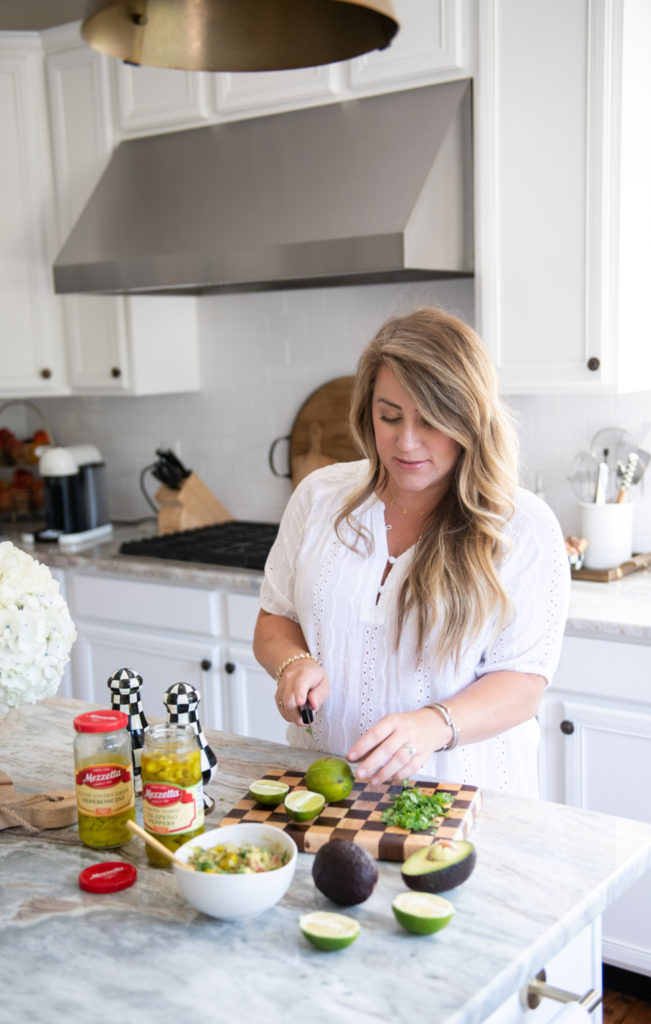Easy Guacamole Recipe by popular Ohio lifestyle blog, Coffee Beans and Bobby Pins: image of a woman slicking a lime on a cutting board next to a couple jars of Mezzetta jalapeno peppers, avocados, and a bowl of guacamole. 