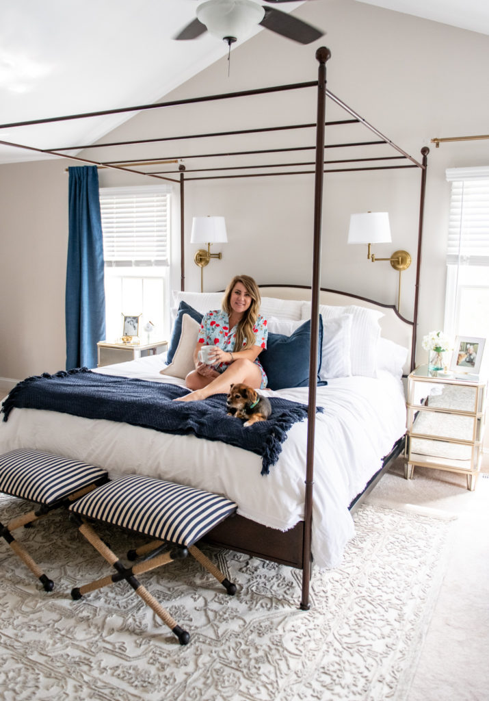 Master Bedroom Remodel Ideas by popular Ohio lifestyle blog, Coffee Beans and Bobby Pins: image of a master bedroom with a canopy bed, tufted ottoman, blue curtains, gold curtain rods, white and blue bedding, gold sconces, and blue and white striped bench. 