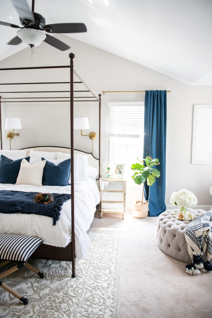 Master Bedroom Remodel Ideas by popular Ohio lifestyle blog, Coffee Beans and Bobby Pins: image of a master bedroom with a canopy bed, tufted ottoman, blue curtains, gold curtain rods, white and blue bedding, gold sconces, and blue and white striped bench. 