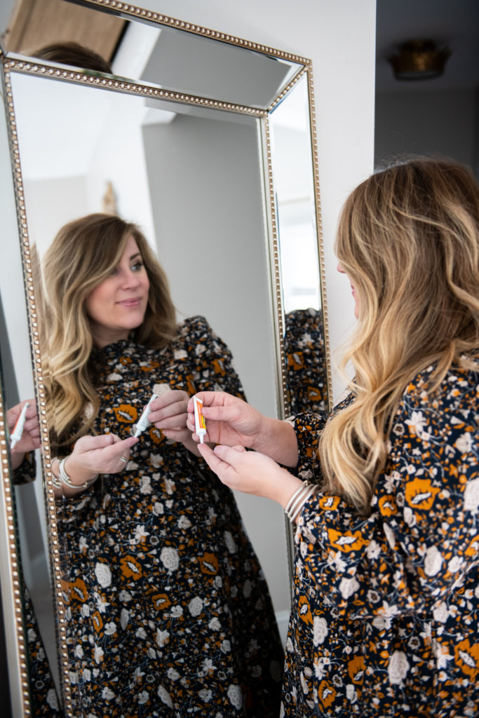 A person standing in front of a mirror posing for the cameraDescription automatically generated |Campho Phenique by popular Ohio lifestyle blog, Coffee Beans and Bobby Pins: image of a woman wearing a black floral dress and looking at herself in a mirror while holding a bottle of Campho Phenique. 