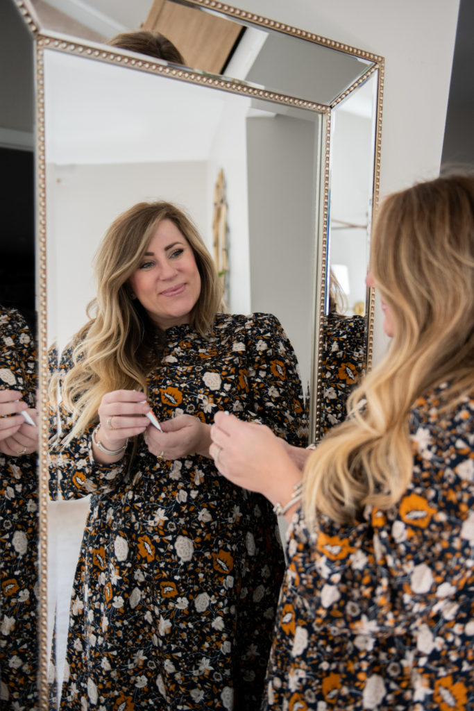 Campho Phenique by popular Ohio lifestyle blog, Coffee Beans and Bobby Pins: image of a woman wearing a black floral dress and looking at herself in a mirror while holding a bottle of Campho Phenique. 