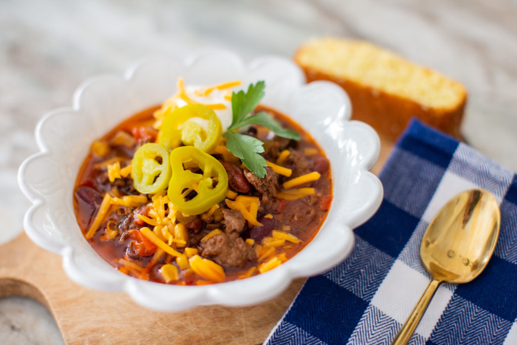 Crockpot Chili by popular Ohio lifestyle blog, Coffee Beans and Bobby Pins: image of some crockpot chili in a white ceramic bowl that's resting on a wooden cutting board next to a slice of cornbread and a blue and white buffalo check cloth napkin with a gold spoon resting on top. 