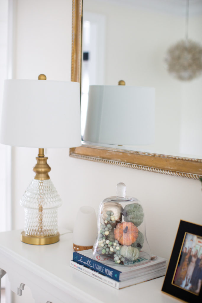 Home Tour by popular Ohio lifestyle blog, Coffee Beans and Bobby Pins: image of a clear glass and gold lamp, black picture frame, wood frame mirror, oil diffuser on a white table. 