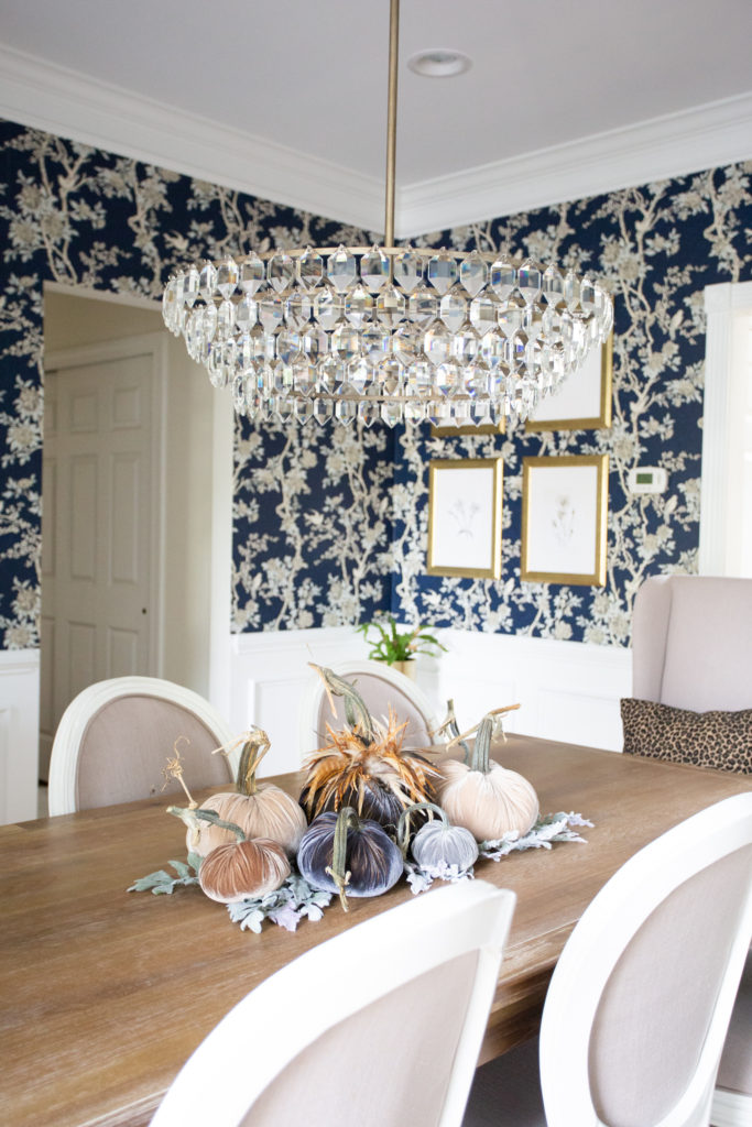 Home Tour by popular Ohio lifestyle blog, Coffee Beans and Bobby Pins: image of a dining room with blue and white floral wallpaper, gold picture frames, modern crystal chandelier, and a farmhouse dining table with a velvet pumpkin centerpiece. 
