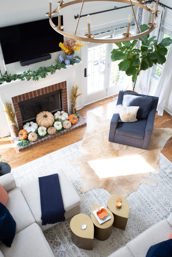 Home Tour by popular Ohio lifestyle blog, Coffee Beans and Bobby Pins: image of a living room decorated with a black side table, vase of flours, cow hide rug, grey couch, blue chair, blue and white area rug. gold chandelier, fiddle leaf fig tree, and faux pumpkins. 