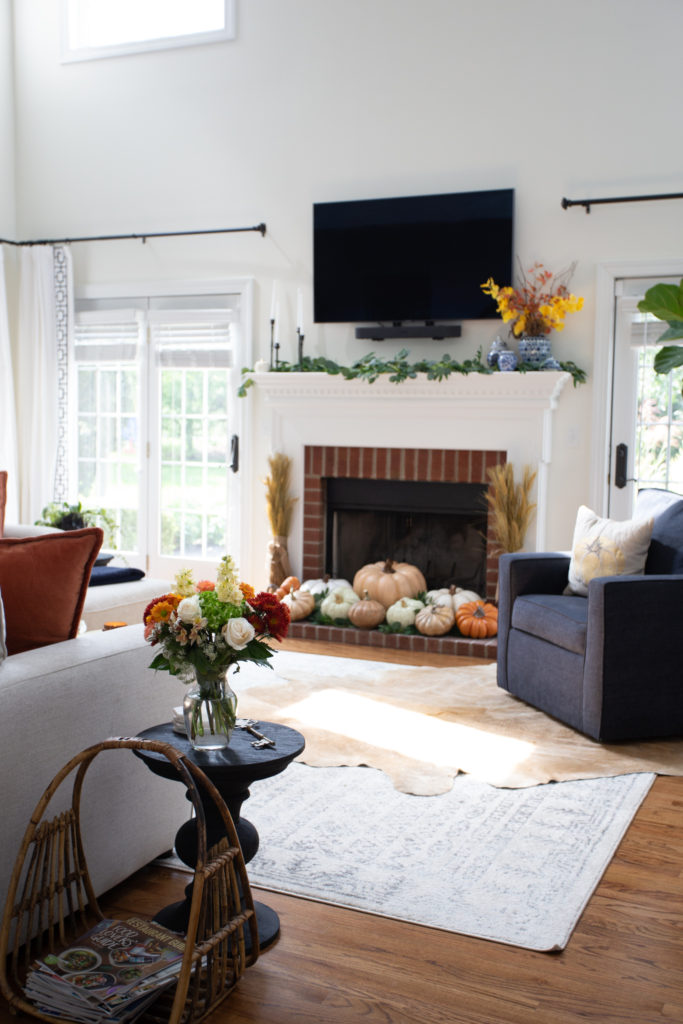 Home Tour by popular Ohio lifestyle blog, Coffee Beans and Bobby Pins: image of a living room decorated with a black side table, vase of flours, cow hide rug, grey couch, blue chair, blue and white area rug. and faux pumpkins. 