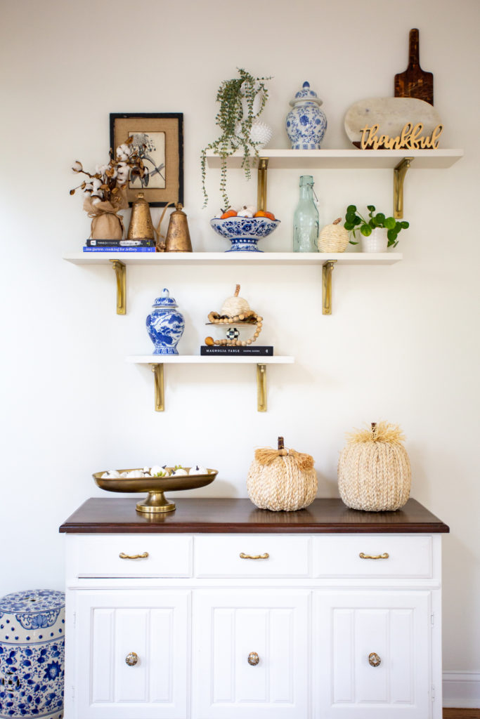 Home Tour by popular Ohio lifestyle blog, Coffee Beans and Bobby Pins: image of a white buffet under white floating shelves decorated with blue and white china, gold cattle bells, house plants, wooden bead garland, straw pumpkins, and books. 