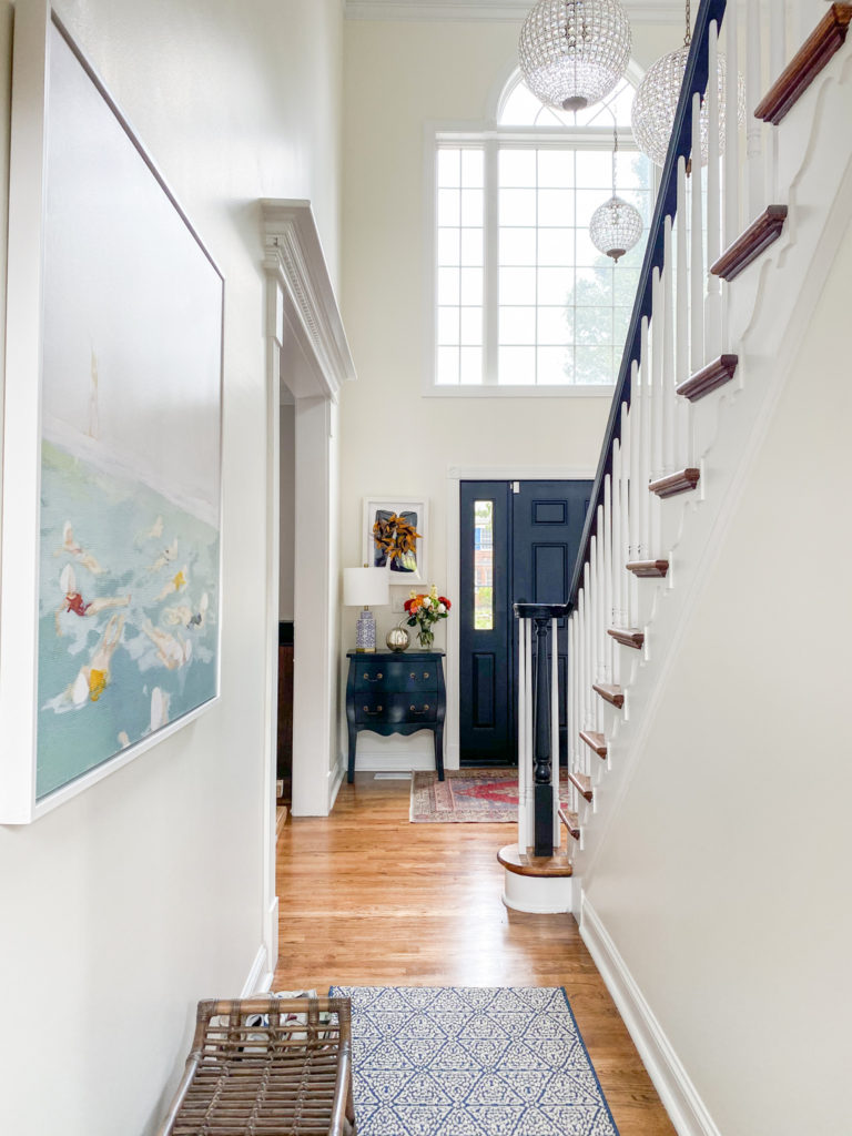 Home Tour by popular Ohio lifestyle blog, Coffee Beans and Bobby Pins: image of a hallway decorated with a bamboo bench, painting of women swimming, blue and white rug runner, black side table, floral arrangement, and silver pumpkin decor item. 