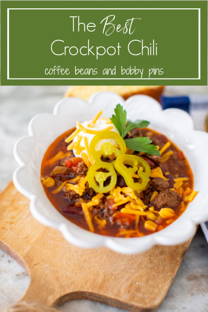 Crockpot Chili by popular Ohio lifestyle blog, Coffee Beans and Bobby Pins: Pinterest image of some crockpot chili in a white ceramic bowl that's resting on a wooden cutting board next to a slice of cornbread and a blue and white buffalo check cloth napkin with a gold spoon resting on top. 