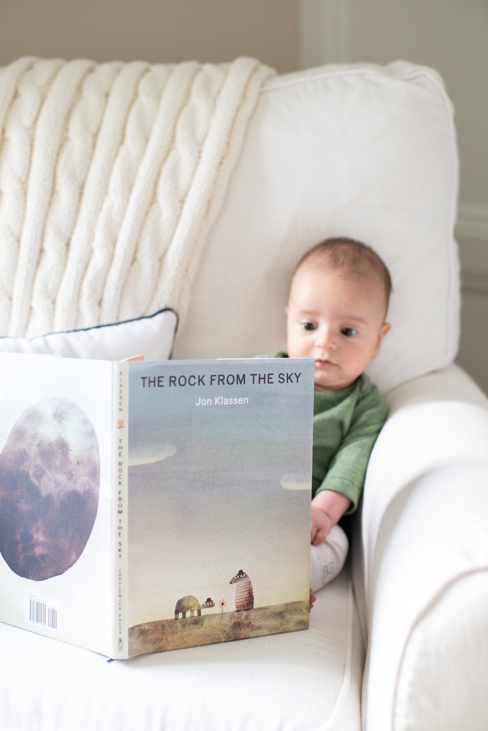 Mommy and Me by popular Ohio motherhood blog, Coffee Beans and Bobby Pins: image of a baby boy sitting on a white armchair and looking at the book The Rock From the Sky. 