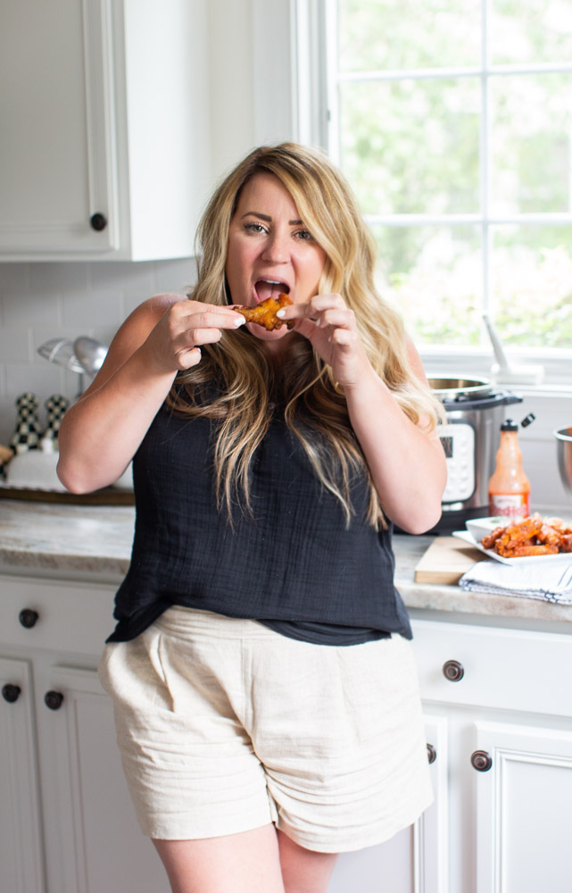 Air Fried Chicken Wings by popular Ohio lifestyle blog, Coffee Beans and Bobby Pins: image of a woman wearing a black tank top and tan shorts and eating a chicken wing in her kitchen. 