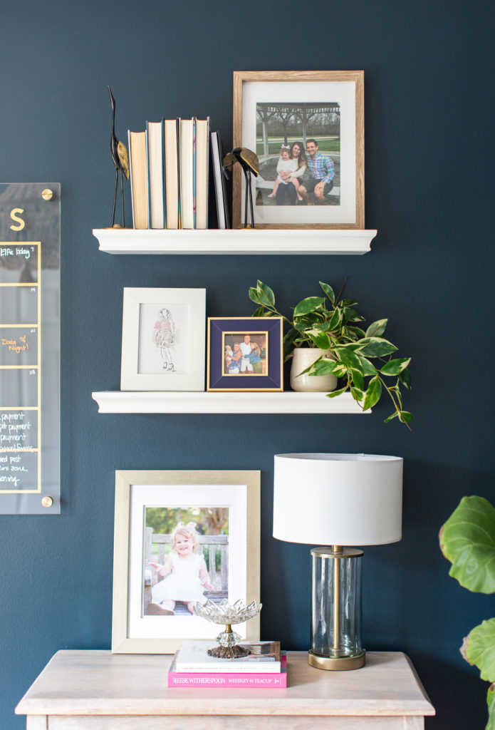 Office Revamp by popular Ohio life and style blog, Coffee Beans and Bobby Pins: image of white floating shelves, books, gold, white and wood picture frames, metal bird decor, indoor plant in a gold pot, and lamp with clear glass base and white shade.
